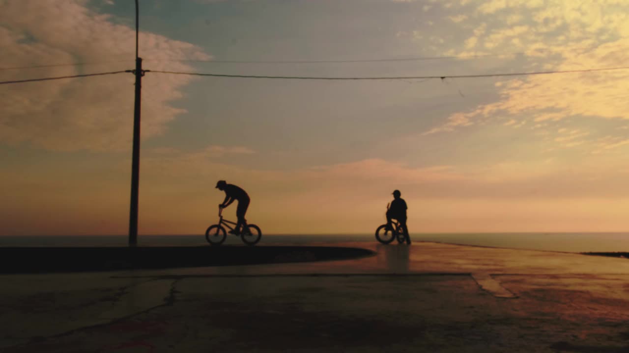 年轻男子在日落时分骑着小轮自行车做坡道跳跃特技。一个骑小轮自行车的人的轮廓无法辨认。BMX骑手。视频下载