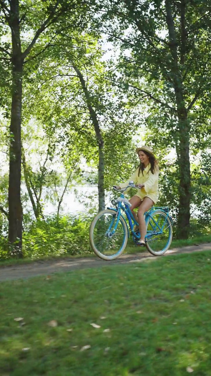竖屏:可爱的年轻女子戴着夏天的帽子，悠闲地骑着蓝色的自行车视频下载
