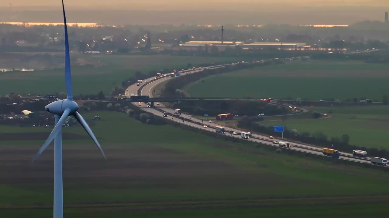 日出时风力涡轮机的超远摄镜头。长焦距在风力涡轮机和附近的高速公路之间建立了连接。早上交通高峰期。视频素材