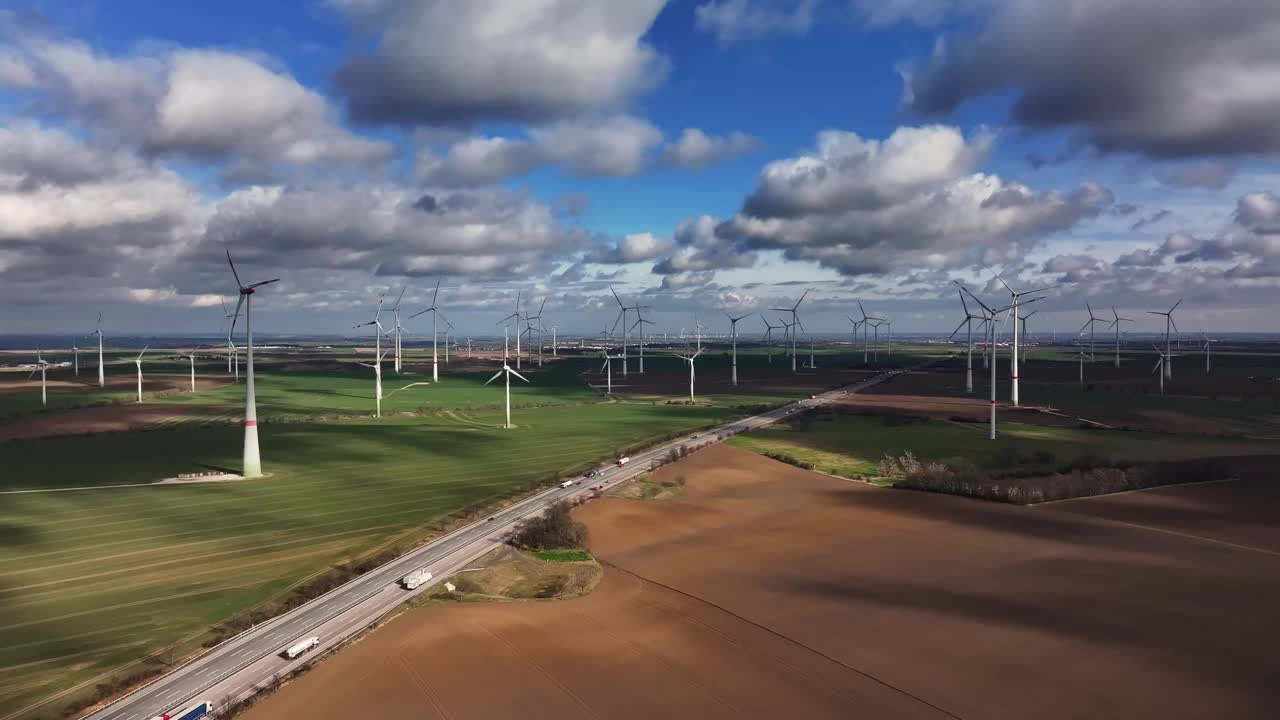沿着高速公路飞行。大型风力发电场分布在几块田地上。阳光下的风力涡轮机。晴朗的一天，有几朵云。公路旁的荒地。视频下载