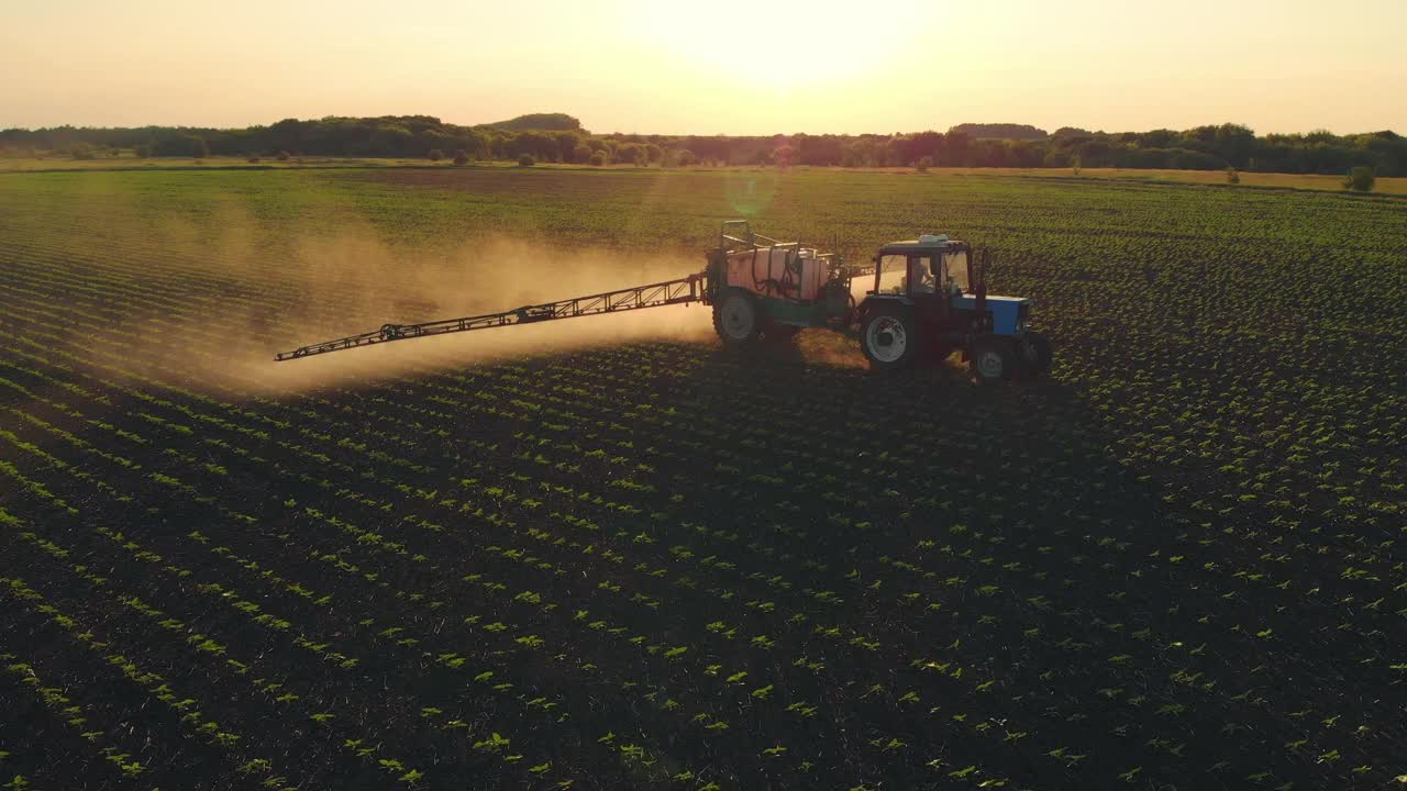 空中镜头。日落时分，农药喷雾器拖拉机在一大片绿色的田野上工作。航拍照片，旁边是一辆拖拉机在麦田里喷洒防病剂。农民产业概念。视频下载