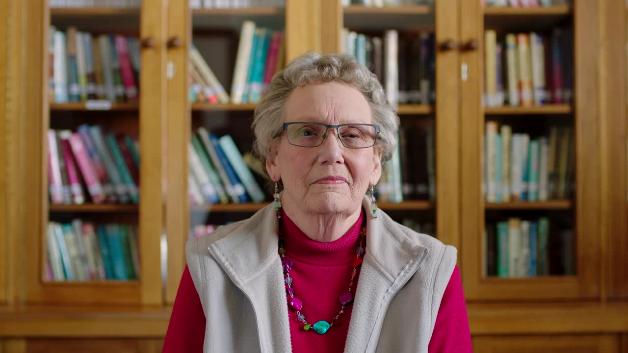 一位年长的女性坐在图书馆里看书，从书架上看小说或文学作品。在教育、学习和研究书店工作的老年图书管理员或教授。视频素材