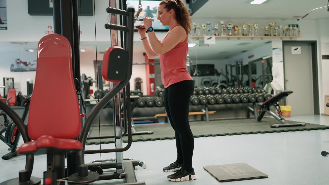 健身房里穿着运动服跪在下拉式举重机上锻炼的健康年轻女子侧视图视频下载