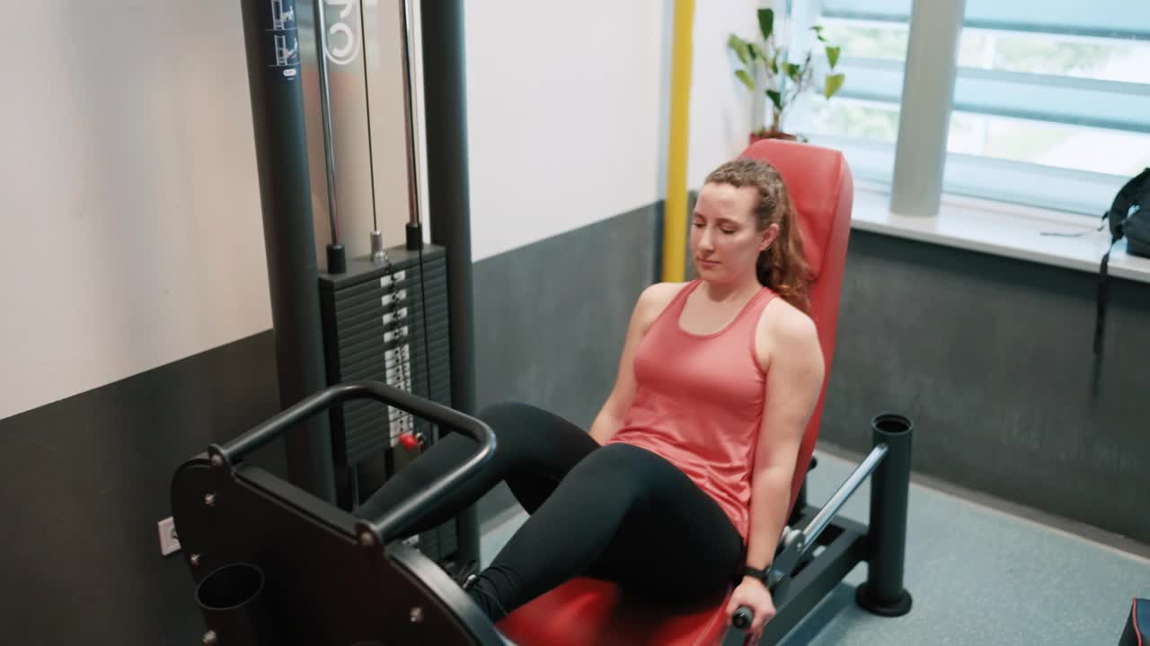 运动的年轻女子在健身房的训练器械上进行腿部按压练习视频下载