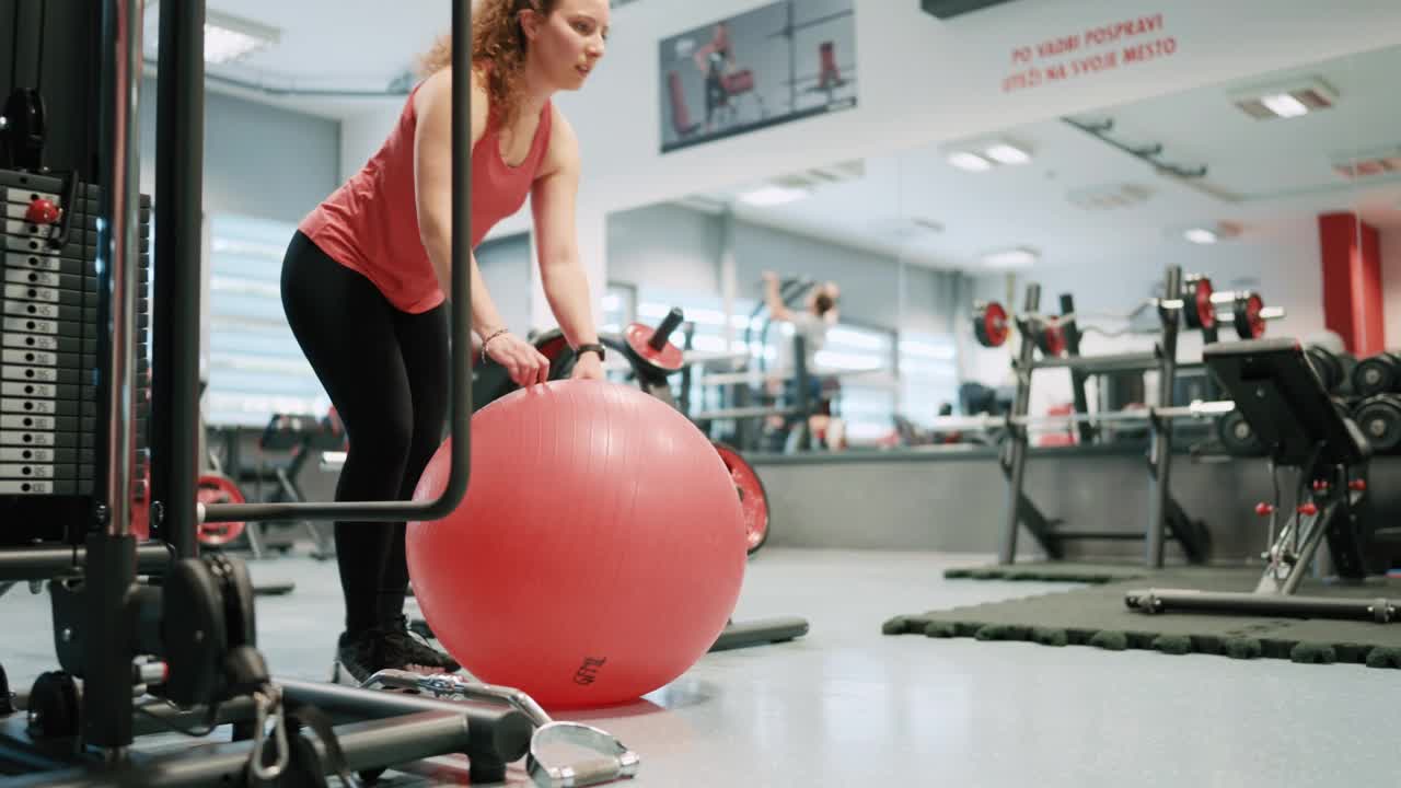 手持拍摄的运动年轻女子穿着运动服，健身球在健身房地板上锻炼视频下载