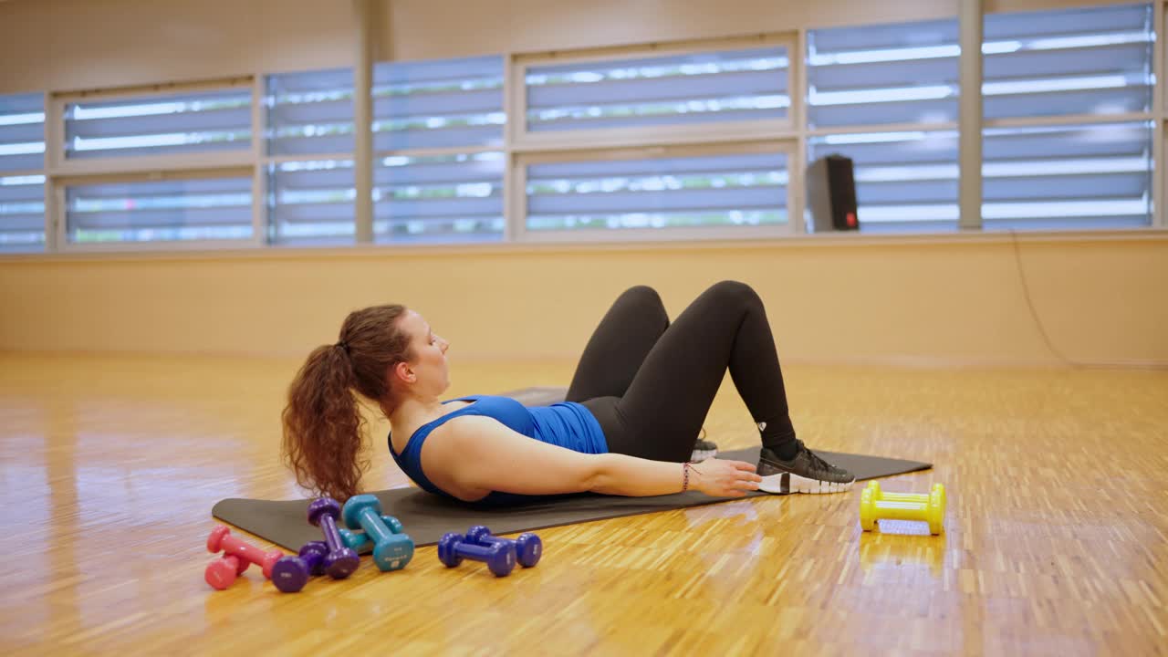 在宽敞健身俱乐部，穿着运动服的年轻女子躺在垫子上做伸展运动的全身侧视图视频下载