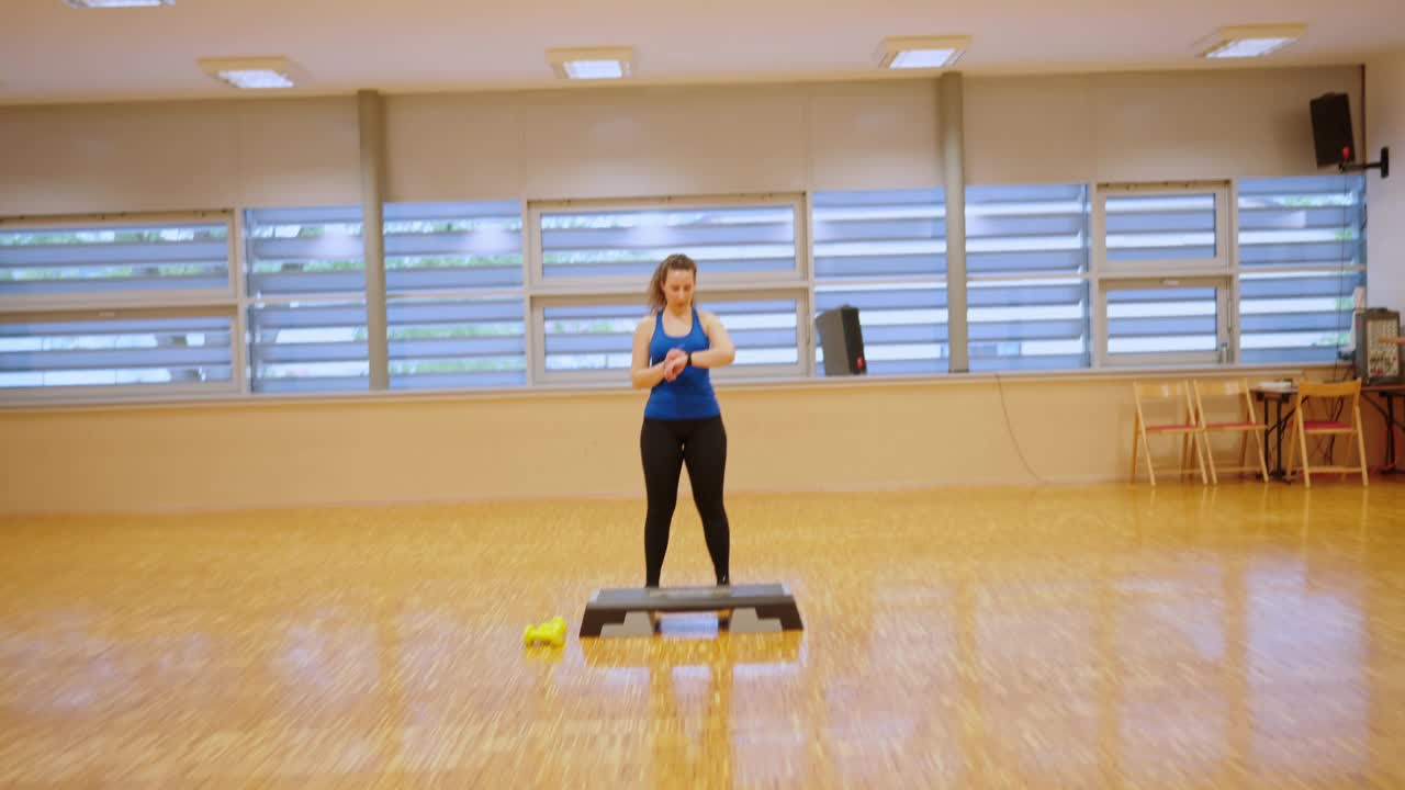 手持拍摄的健康年轻女子穿着运动服，使用健身追踪器，在健身俱乐部做踏步运动视频下载