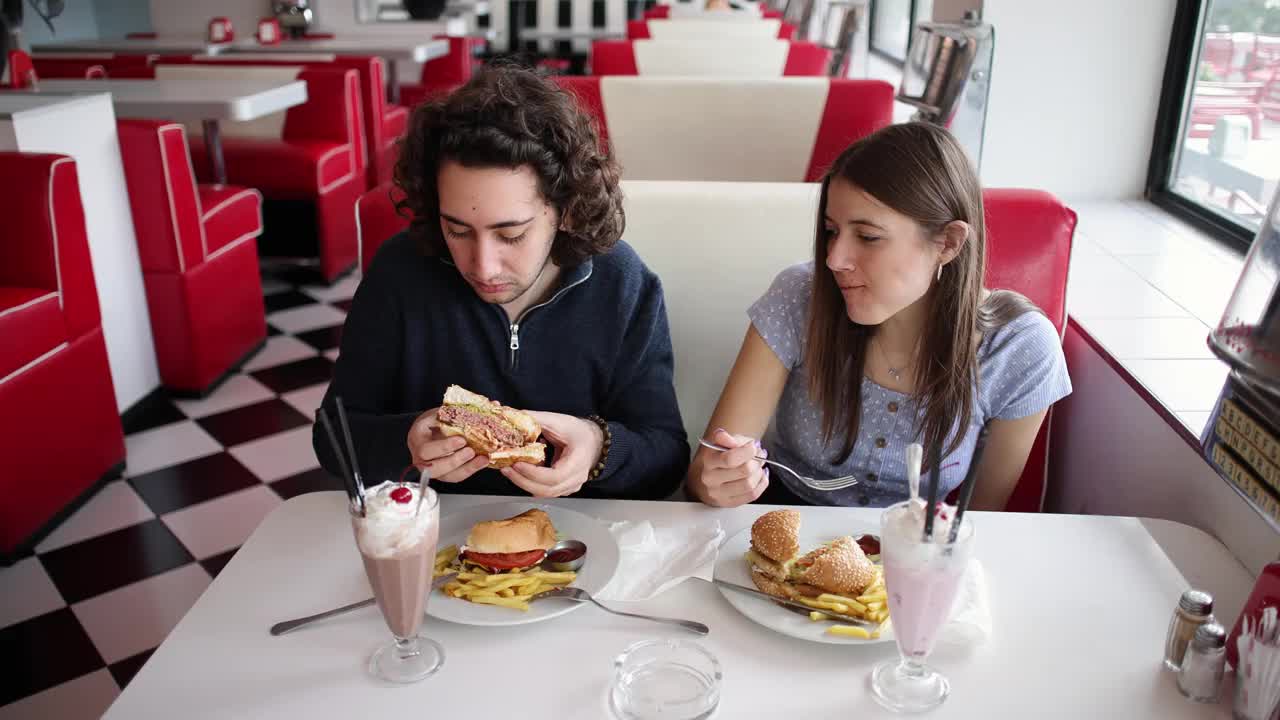 现代年轻的白人夫妇在复古的快餐店里吃汉堡和奶昔视频下载