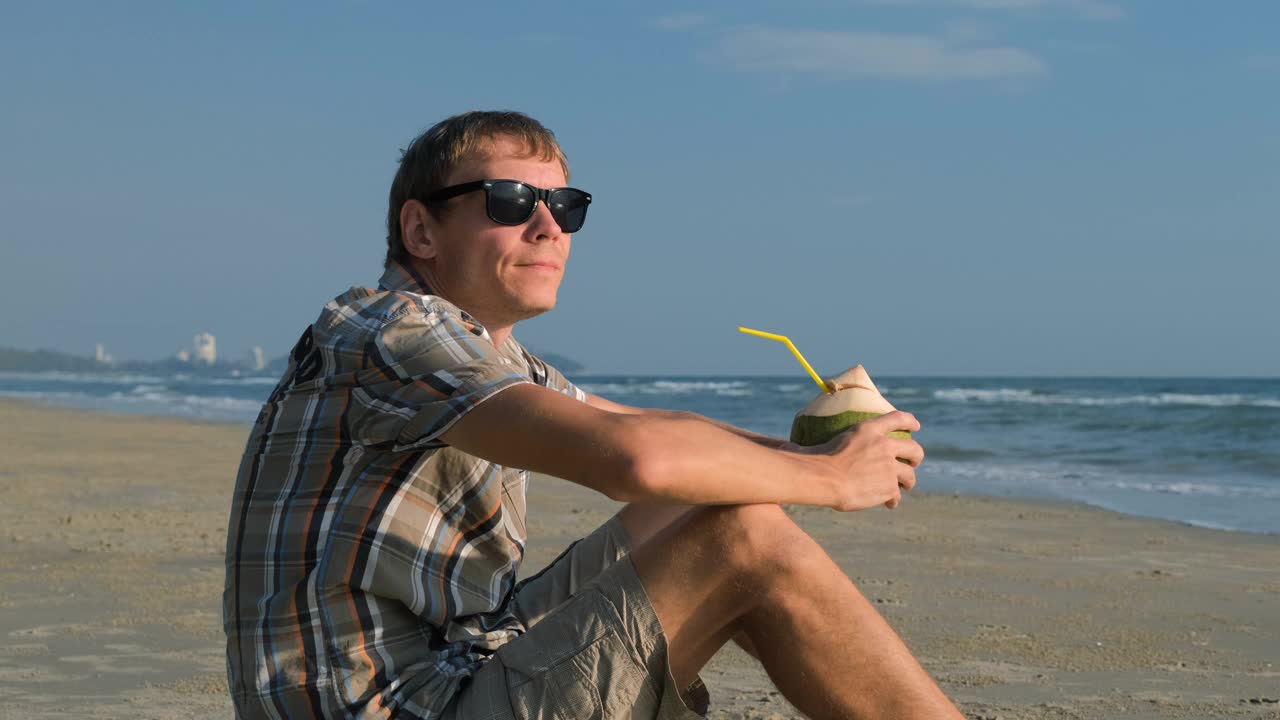 戴墨镜的男子在沙滩上喝椰子汁视频下载