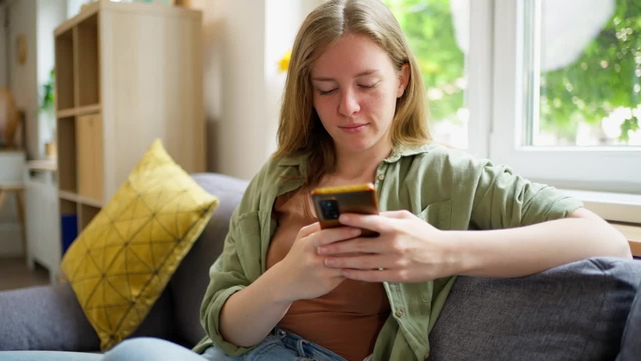 一个年轻的白人女人坐在沙发上玩手机视频素材