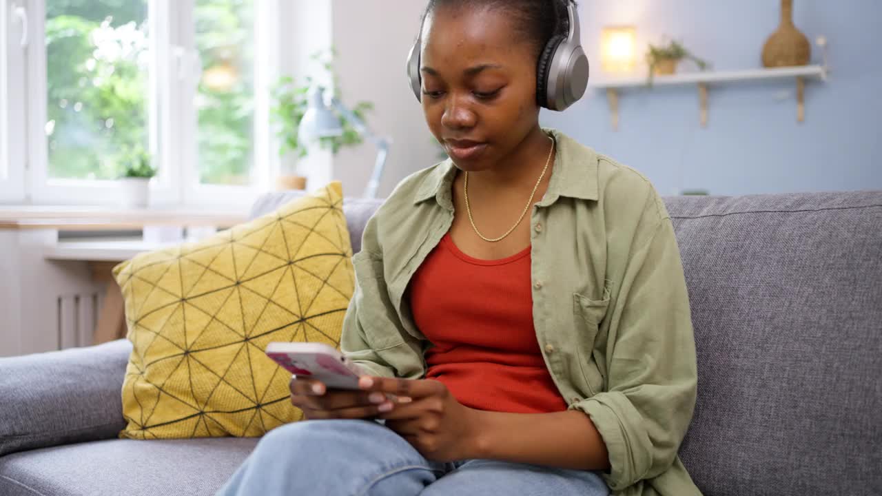 无忧无虑的黑人妇女坐在沙发上，一边听着无线耳机上的音乐一边跳舞视频素材