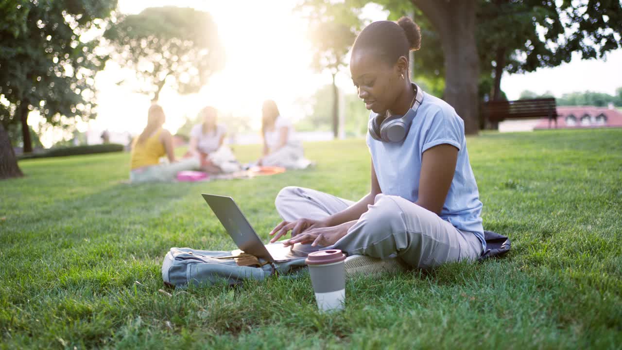 一个年轻的黑人女性的肖像，坐在草地上使用笔记本电脑工作或学习视频下载