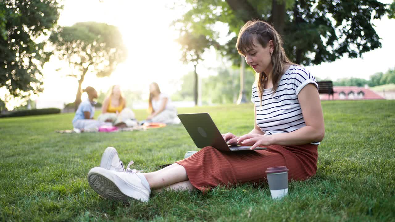 一位年轻的白人女性坐在草地上，使用笔记本电脑工作或学习视频下载