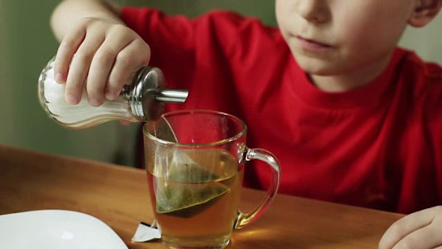 小男孩正在把糖倒进一杯茶里，并用勺子搅拌。视频素材