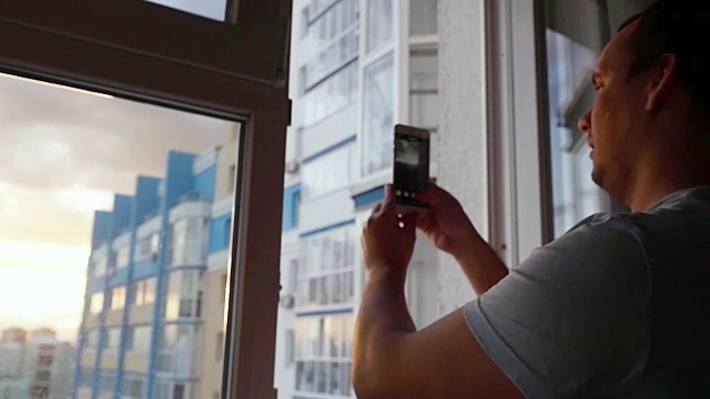一个年轻人在惊人的日落中用手机用玻璃窗户在城市上空拍摄了一幅慢动作的照片视频下载