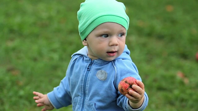漂亮的宝宝在家带着苹果在花园里散步。戴着帽子，穿着毛衣，背景是草地的孩子视频下载