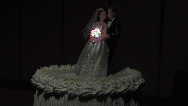 蛋糕上的新婚夫妇小雕像和一束明亮的花束在黑暗中闪闪发光。视频下载