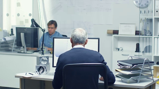 在繁忙的建筑工程局高级工程师的Backview与3D模型在他的个人电脑上工作。也可以看到他的助手在他的办公桌上工作。视频素材
