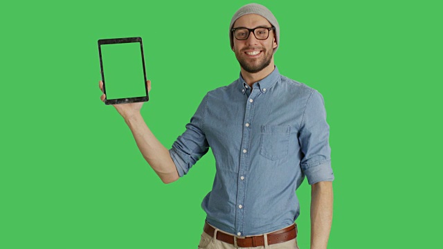 中景一名戴着帽子和眼镜的时尚男子用一只手拿着平板电脑，用另一只手做滑动触摸手势。平板电脑和背景都是绿屏。视频下载