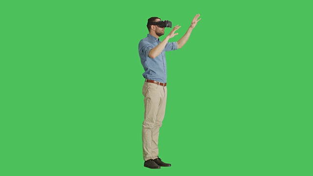 一个英俊的男人戴着虚拟现实眼镜与外部世界互动的长镜头，而相机在他周围旋转。背景为绿幕。视频下载