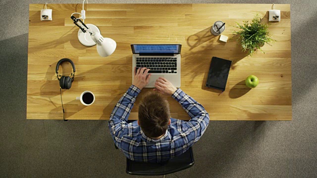俯视图的一个年轻的创造性的人在他的笔记本电脑上工作，而坐在他的木制书桌。桌子上也有:咖啡杯，智能手机，笔记本电脑，台灯，植物。视频素材