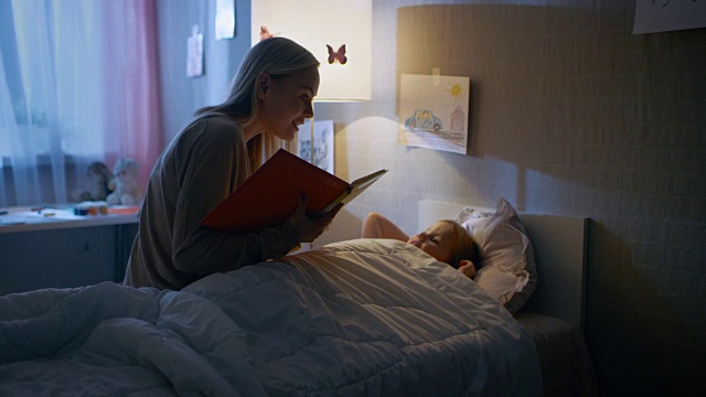 年轻慈爱的母亲读睡前故事给她美丽的小女儿，她在她的床上睡觉。视频购买