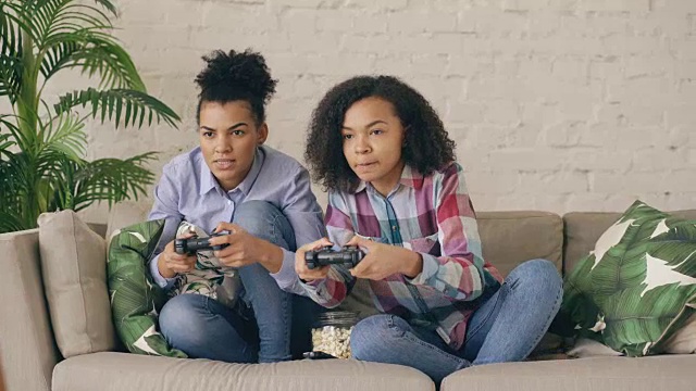 两个混合种族卷曲的女孩朋友坐在沙发上玩控制台电脑游戏与手柄，有乐趣在家里视频下载