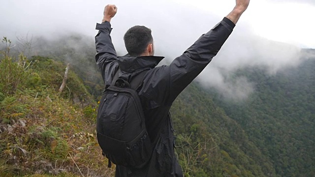 一名年轻男性徒步旅行者背着背包爬上山顶，举起双手。一名游客站在美丽的峡谷边缘，胜利地伸出双臂。慢镜头后视图视频下载