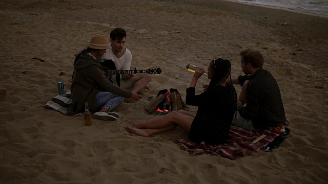 年轻的朋友们坐在沙滩上的火堆旁，烤着香肠，喝着啤酒，弹着吉他。Slowmotion拍摄视频素材