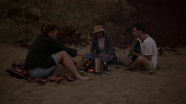 年轻的朋友们晚上坐在海滩上的火边，烤着香肠，喝着啤酒，弹着吉他。Slowmotion拍摄视频素材