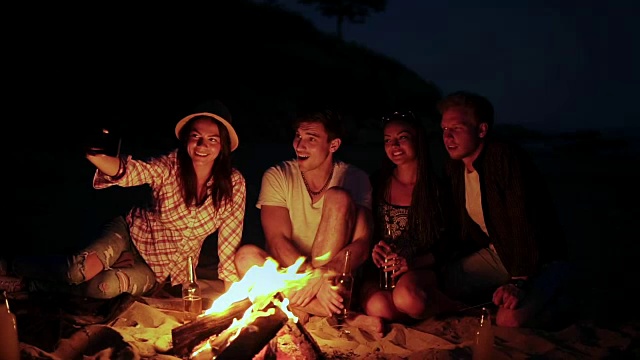 年轻人晚上在海滩上用篝火野餐。愉快的朋友在电话里拍照。Slowmotion拍摄视频素材