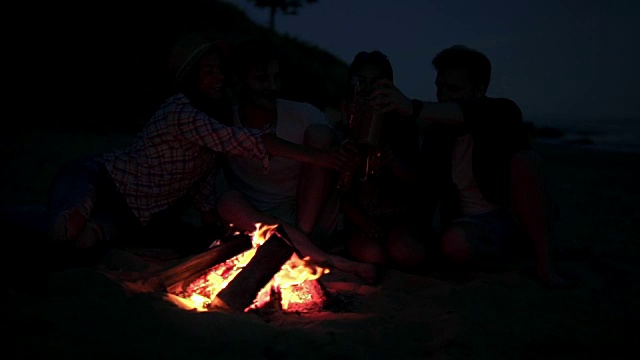晚上，年轻人在海滩上篝火野餐，喝啤酒，欢呼。快乐的朋友们唱歌弹吉他。Slowmotion拍摄视频素材