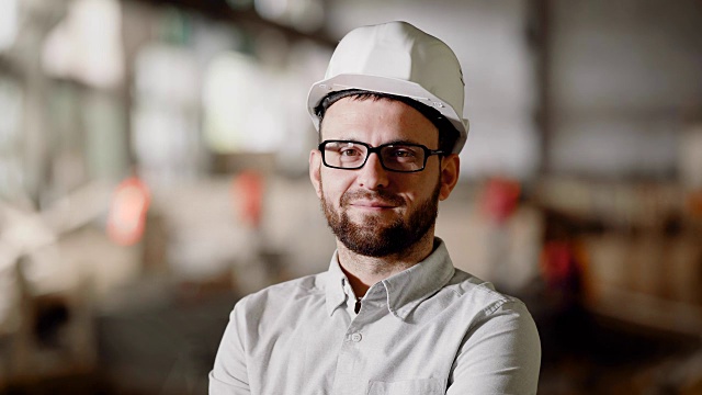 近距离的胡子男子在白衬衫，保护头盔和眼镜站在施工区域。在建筑工地工作的年轻建筑师。男工程师控制建造过程视频下载