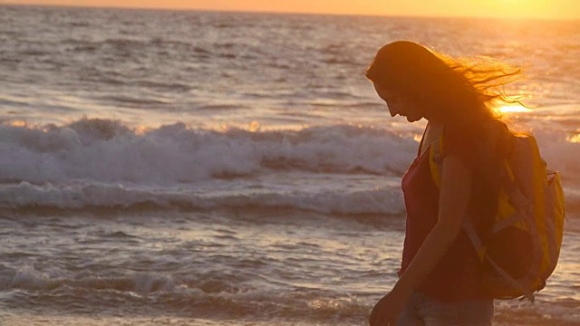 年轻的女性游客背包沿着海滩在日落。日出时分，美丽的年轻女旅行者踏上了海岸。在旅行或暑假期间徒步的女孩。慢动作视频购买