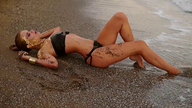 性感苗条的模特在黑色皮革比基尼躺在海滩上的海浪。迷人的年轻女子与专业艺术金色的化妆和马尾辫在沙滩上的海边。Slowmotion拍摄视频下载