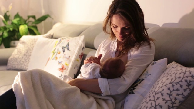 年轻美丽的母亲，在昏暗的灯光下给她的新生儿喂奶。妈妈母乳喂养婴儿视频下载