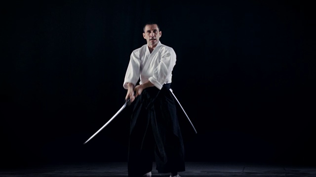 合气道大师穿着传统的日本武士袴衣服拿出他的日本剑鞘和秋千。他在聚光灯下，黑暗包围着他。拍摄孤立的黑色背景。视频下载