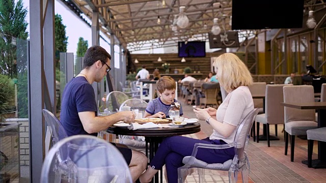 一个年轻的家庭在一家餐馆用餐，一个女人被她的丈夫拍了下来，她正在喝茶，他们的孩子用吸管喝奶昔视频素材