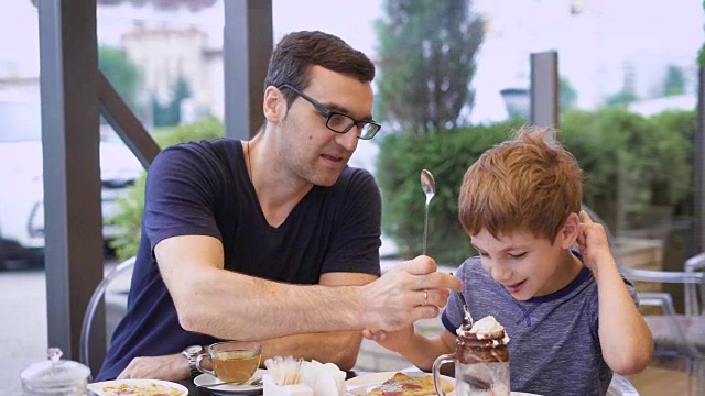 一位年轻的父亲尝试他儿子的牛奶鸡尾酒与冰淇淋和巧克力，儿子笑了，一家人正在餐厅用餐视频素材