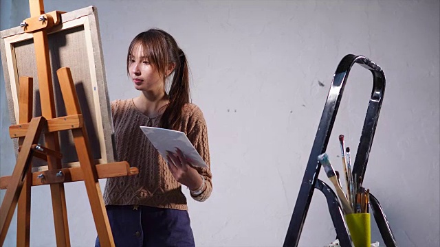 年轻女子在有大窗户的艺术空间的帆布上画油画视频下载