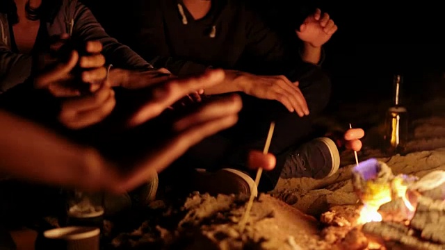 野餐的年轻人坐在篝火在海滩和温暖他们的手在晚上。特写视图视频素材
