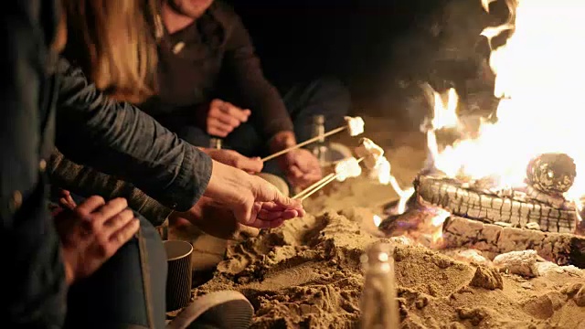 一群年轻的多民族的人坐在篝火上的海滩深夜，在火上烤棉花糖棒在一起视频素材