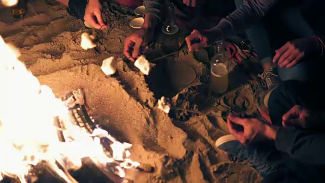 从上往下看:双手握着棉花糖棒，在晚上炸棉花糖。深夜一群人坐在火旁视频素材