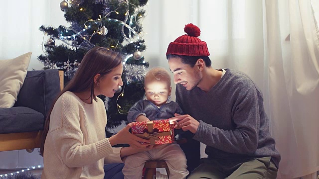 快乐的家庭和宝宝带着圣诞礼物在家里的圣诞树旁。孩子打开礼品盒视频下载