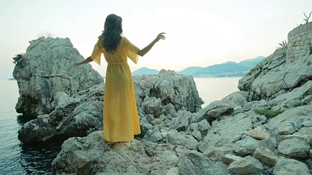 夏日的傍晚，年轻的黑发女郎站在海边的岩石上移动着身体视频下载