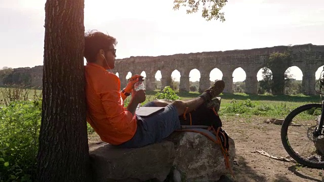 骑自行车的人坐在树下的一块岩石上，用他的笔记本电脑听音乐，在古罗马的渡槽前喝水视频下载
