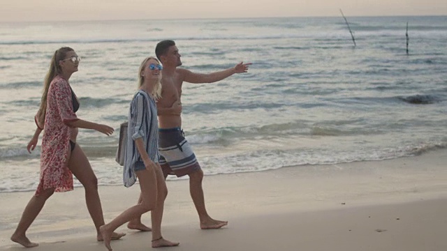 两个美丽的女人和英俊的健康的男人沿着海滩散步，聊天和玩。美丽的风景海在背景。视频购买
