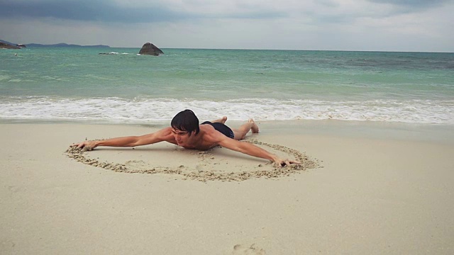年轻人躺在沙滩上，在海边潮湿的沙滩上画了一个心形符号。缓慢的运动。1920 x1080视频下载