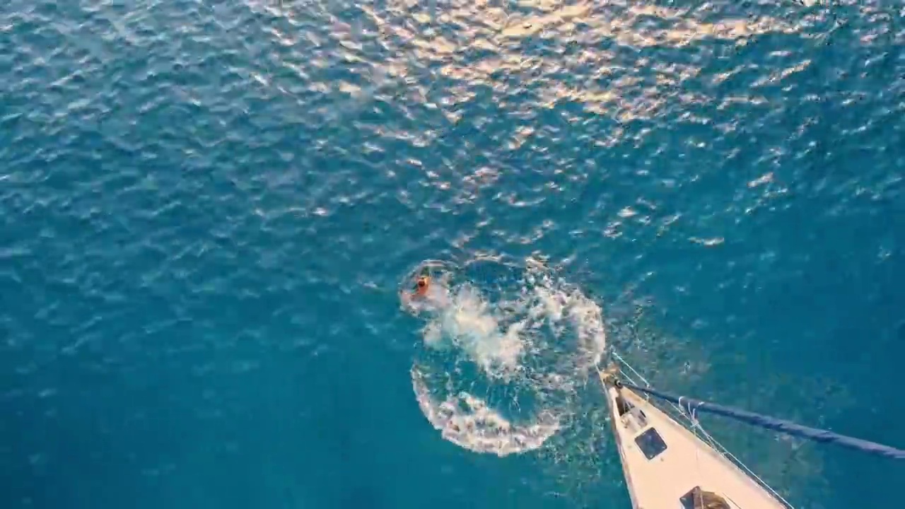 4K无人机实时拍摄男子从帆船上跳向阳光明媚的蓝色海洋视频下载
