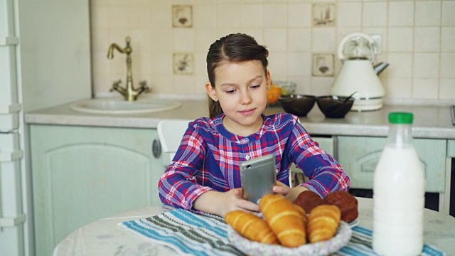 美丽的小女孩早上坐在厨房的桌子边玩智能手机。童年、人、技术的概念视频素材